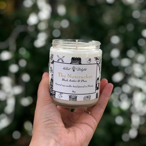 May include: A white candle jar with a black and white illustration of a nutcracker, a Christmas tree, and other holiday imagery. The label reads "Nolent Delights The Nutcracker Black Amber & Plum Natural soy candles hand poured in Florida 9oz".