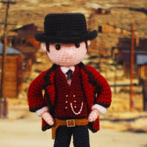 Op de afbeelding: Een gehaakte pop verkleed als cowboy, met een zwarte hoge hoed, een rood jasje met zwarte randen, een wit overhemd met een zwarte das en een donkere broek. De figuur heeft een bruine riem met een holster en een klein zilveren kettinkje.