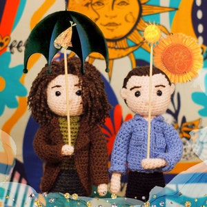 Puede incluir: Dos muñecas de crochet, una sosteniendo un paraguas marrón y la otra una flor amarilla, se encuentran frente a un fondo colorido.