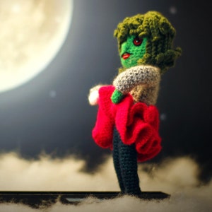 May include: A green crocheted doll with red and white clothing stands in front of a large, bright moon. The doll has a green face with red eyes and a crocheted hairstyle.