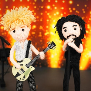 May include: Two crocheted dolls, one playing a guitar and the other holding a microphone, are performing on a stage with a sparkly orange backdrop.