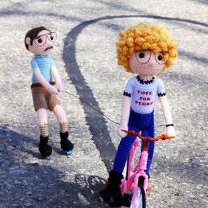Könnte beinhalten: Gehäkelte Puppen, die Charaktere darstellen, eine auf einem rosa Fahrrad und die andere auf Rollschuhen. Die Puppe auf dem Fahrrad trägt ein T-Shirt mit der Aufschrift "Vote for Pedro". Beide Puppen tragen eine Brille und befinden sich auf einer gepflasterten Oberfläche.