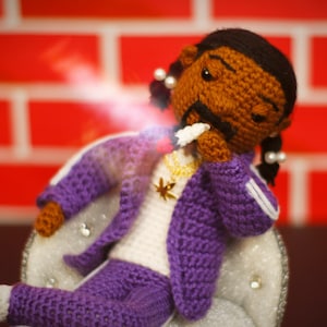 May include: A crocheted doll with brown skin, purple clothing, and white accents sits on a white chair with rhinestones. The doll has black hair in pigtails with white beads. The doll is holding a white object in its hand.