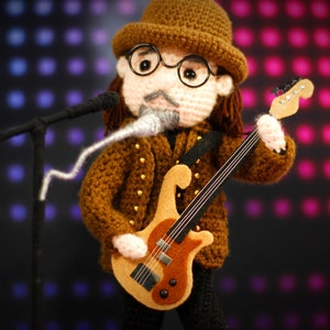 May include: A crocheted musician doll wearing a brown hat and glasses, playing a light brown and orange guitar. The doll has a mustache and wears a brown jacket with gold buttons. It stands before a microphone on a black stand.