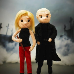 May include: Two crocheted dolls, one with blonde hair wearing a black dress and red trousers, holding a wooden stick, and the other with short blonde hair wearing a black coat and black trousers.