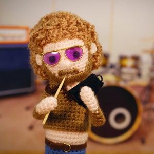 Op de afbeelding: Een gehaakte pop met bruin krullend haar, die een gouden bril met paarse glazen draagt, een bruine trui en een blauwe spijkerbroek. De pop houdt een houten drumstick en een zwart notitieboek vast.