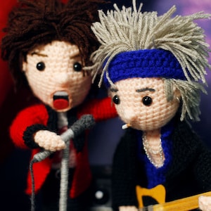 Puede incluir: Muñecos de ganchillo de dos músicos. Uno canta a un micrófono, con una chaqueta roja. El otro toca una guitarra amarilla, con una cinta azul en la cabeza. Ambos muñecos tienen rasgos faciales detallados y pelo de hilo.