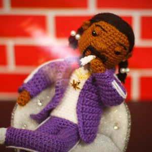 May include: A crocheted doll wearing a purple tracksuit with white accents, a gold chain necklace with a star pendant, and a white cigarette in its mouth. The doll has brown skin, black hair, and a serious expression.
