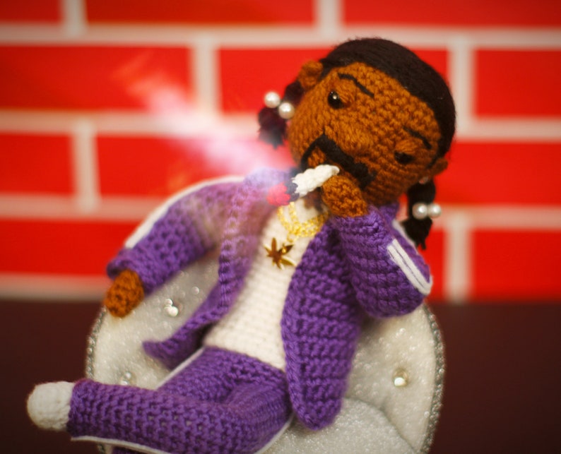 May include: A crocheted doll wearing a purple tracksuit with white accents, a gold chain necklace with a star pendant, and a white cigarette in its mouth. The doll has brown skin, black hair, and a serious expression.