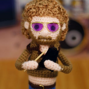 Pode incluir: Uma boneca de croch&ecirc; com cabelo castanho encaracolado, vestindo roupas marrons, &oacute;culos de aro dourado com lentes roxas, e segurando um pequeno objeto preto e um pau de madeira.