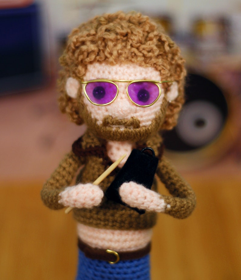 Op de afbeelding: Een gehaakte pop met bruin haar, een bruin shirt, blauwe jeans en gouden bril met paarse glazen. De pop houdt een klein zwart tamboerijn en een houten stok vast.