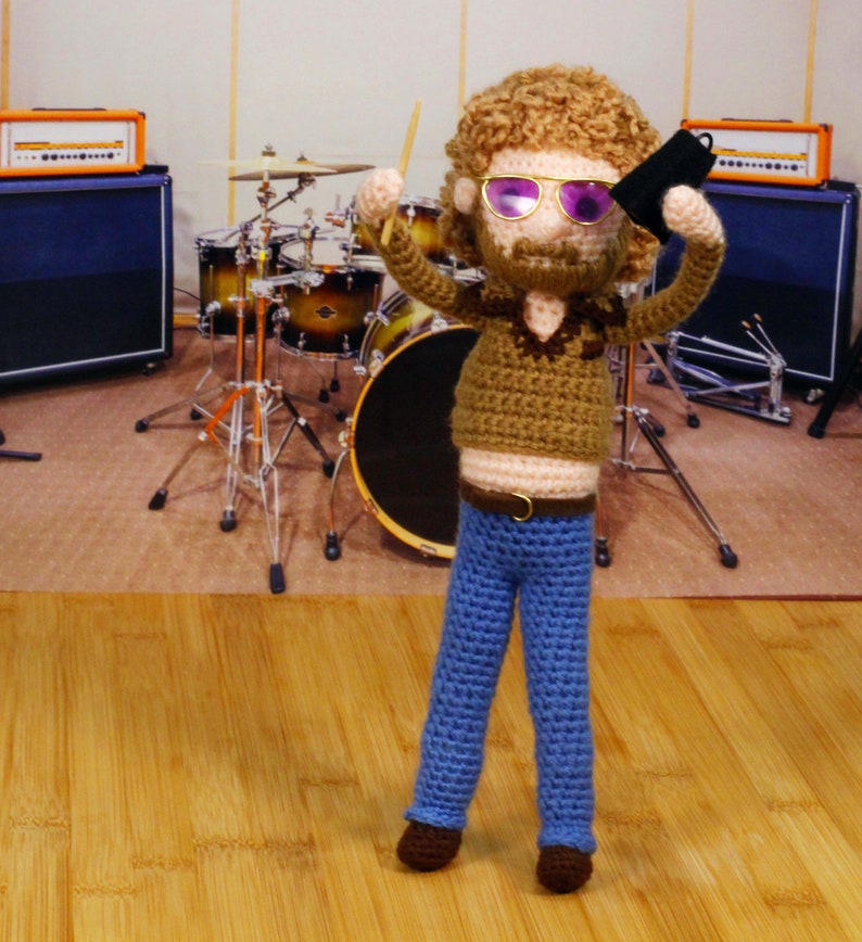 Pode incluir: Uma boneca de croch&ecirc; vestindo jeans azul, uma camisa marrom e &oacute;culos de sol, segurando uma baqueta e um objeto preto, na frente de um kit de bateria com amplificadores laranja.