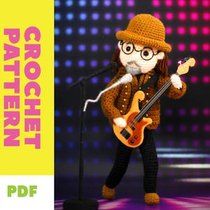 May include: A crocheted musician doll with glasses, a hat, and a guitar, stands in front of a microphone. The doll wears a brown jacket and black pants. The words "CROCHET PATTERN" and "PDF" are displayed on the left side of the image.