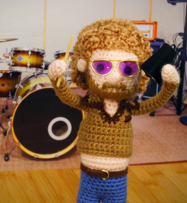 Op de afbeelding: Een gehaakte pop die een bruine broek, een bruin shirt met een bruin en wit ontwerp en een blauwe spijkerbroek draagt. De pop heeft krullend bruin haar, een baard en draagt een paarse zonnebril. De pop houdt een drumstick en een zwarte microfoon vast.