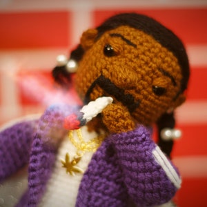 May include: A crocheted doll with brown skin, black hair, and a purple jacket. The doll is holding a white object in its hand and has a gold chain necklace with a marijuana leaf pendant.