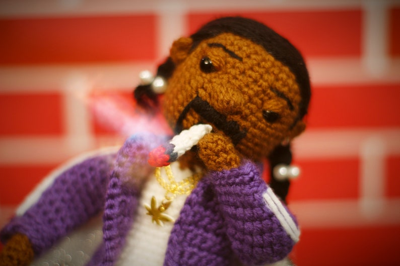 May include: A crocheted doll with brown skin, black hair, and a purple jacket. The doll is holding a white object in its hand and has a gold chain necklace with a marijuana leaf pendant.