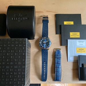 May include: A blue and gold wristwatch with a black leather band and a black box. The watch has a blue face with white markings and a date window. The box has the word "Breitling" printed on it.