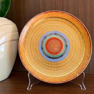 May include: A decorative ceramic plate with a gold, red, blue, and green concentric circle design. The plate has a floral pattern around the edge.