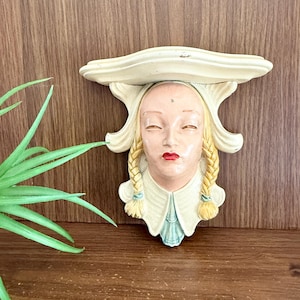 May include: A vintage wall shelf featuring a painted face with closed eyes, red lips, and blonde braids. The shelf is off-white with a decorative design, mounted on a wooden surface. The shelf is a unique decorative item.