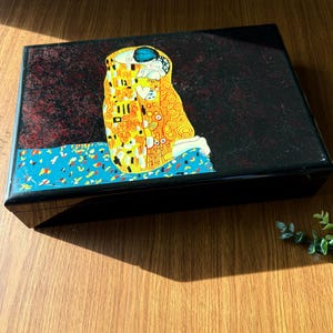 May include: A black rectangular box with a glossy finish. The lid features a reproduction of Gustav Klimt's "The Kiss" painting, with gold, yellow, and blue accents. The box sits on a wooden surface.
