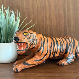 May include: Orange and black tiger figurine with an open mouth, showing its teeth. The tiger is lying down, with its front paws extended. The figurine is on a wooden surface, with a potted plant in the background.