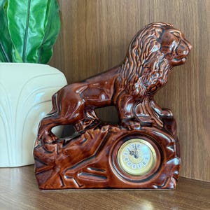 May include: A brown ceramic lion figurine with a clock face. The lion is posed atop a rocky base, with a gold-colored clock face featuring Roman numerals. The figurine is a decorative item.