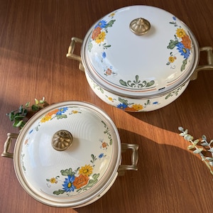 May include: Two white enamel pots with lids, adorned with floral patterns in yellow, blue, and orange. Each pot features brass handles and a brass knob on the lid. The pots are set on a wooden surface, showcasing a vintage aesthetic.