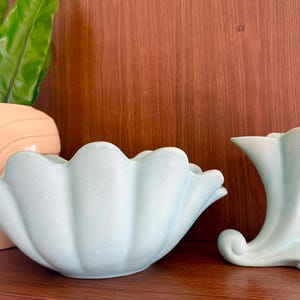 May include: Two light blue ceramic vases. One is a large, scalloped bowl, and the other is a smaller, curved vase with a flared top. The vases are on a wooden surface with a wooden background.