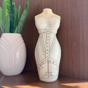 May include: A cream-colored mannequin torso sculpture featuring a hand-drawn black line illustration of a strapless dress. The design includes a lace-up bodice detail, a central row of buttons, and decorative scrollwork patterns along the hemline.