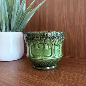 May include: A small, round, dark green ceramic planter featuring a raised, embossed forest motif around the exterior. The planter has a glossy finish and a scalloped base design. It is positioned in front of a white pot containing a green plant.