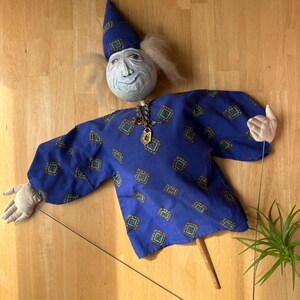 May include: A blue puppet with a white face, a pointed hat, and a gold chain necklace with a key charm. The puppet has a blue and white patterned shirt and is holding a wooden stick.