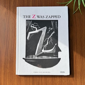 May include: A hardcover book titled "THE Z WAS ZAPPED" by Chris Van Allsburg. The cover features a black and white illustration of a large letter "Z" being struck by lightning, with a stage curtain backdrop.