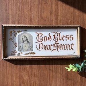 May include: A framed religious sign with the words "God Bless Our Home" in ornate lettering. The sign features an image of Jesus and floral accents in gold. The frame is a light gold color.