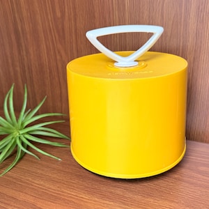 May include: A bright yellow cylindrical container with a smooth finish and a white triangular handle attached to the top center. The container features a flat lid and a solid, opaque body, providing a simple and functional storage solution.