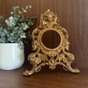 May include: A gold-colored metal picture frame featuring an ornate design with a central circular opening. The frame is decorated with a sculpted cherub at the top and two bearded faces on the sides, supported by two curved feet at the base.