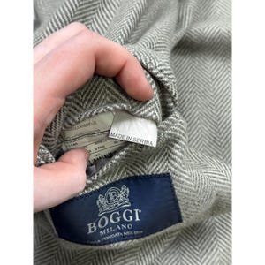 May include: A light gray and white herringbone patterned blazer featuring a dark blue interior label with the text BOGGI MILANO CASA FONDATA NEL 1939. A white tag attached to the inner lining displays the text MADE IN SERBIA along with fabric details.