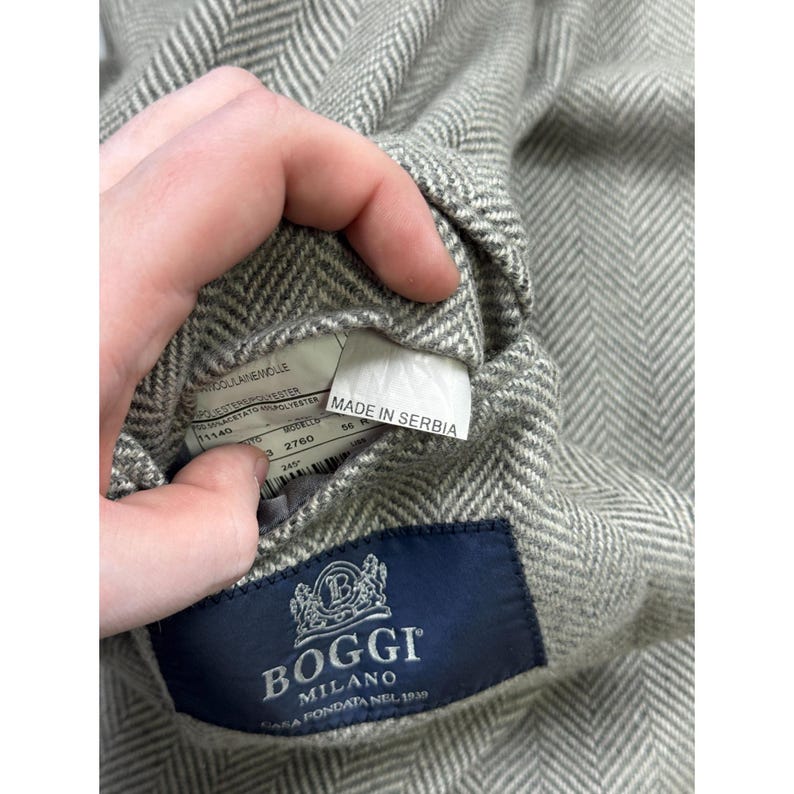 May include: A light gray and white herringbone patterned blazer featuring a dark blue interior label with the text BOGGI MILANO CASA FONDATA NEL 1939. A white tag attached to the inner lining displays the text MADE IN SERBIA along with fabric details.
