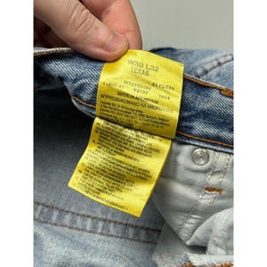 May include: Close-up of a pair of light blue denim jeans with a yellow tag. The tag reads "W30 L32 TEXAS" and includes additional text about the jeans' origin and care instructions. The jeans have a button closure and orange stitching.