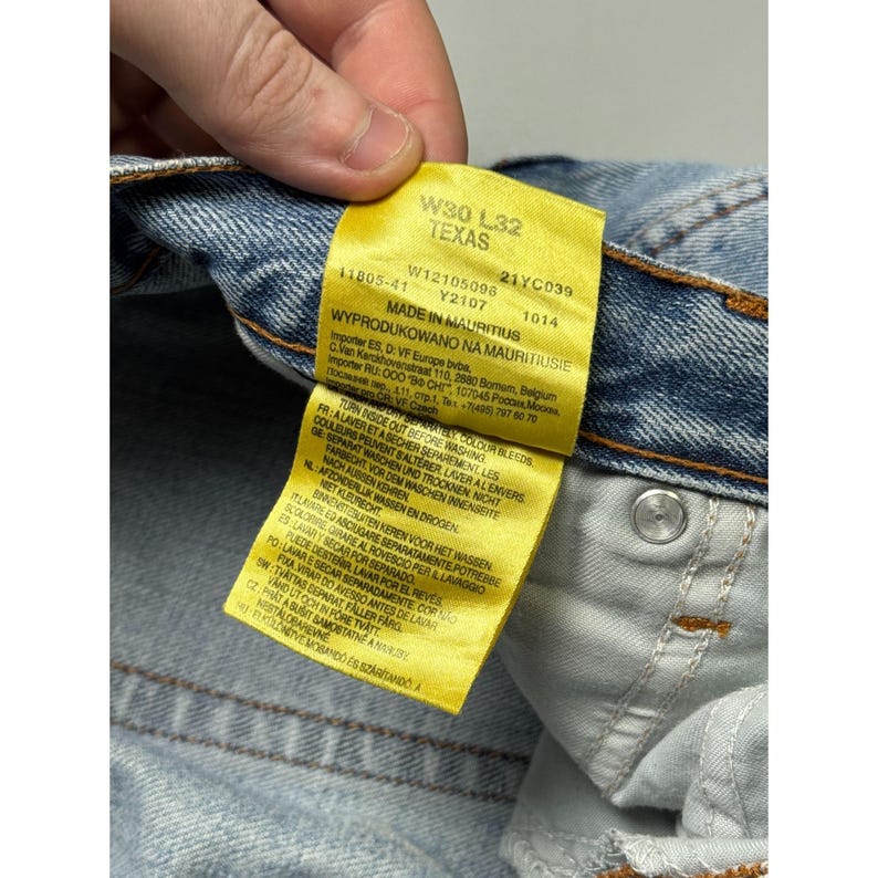 May include: Close-up of a pair of light blue denim jeans with a yellow tag. The tag reads "W30 L32 TEXAS" and includes additional text about the jeans' origin and care instructions. The jeans have a button closure and orange stitching.