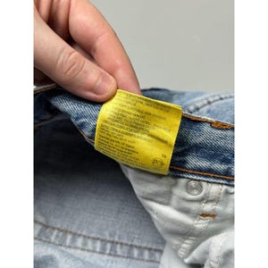 May include: Close-up of light blue denim jeans with a yellow tag. The tag has text in multiple languages. The jeans have a silver button and a white interior pocket.