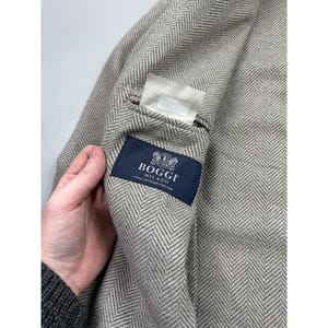 May include: A light beige and white herringbone patterned blazer featuring a dark blue interior label with the white text Boggi Milano Casa Fondata Nel 1939 and a small white rectangular tag positioned above the brand label on the inner lining.
