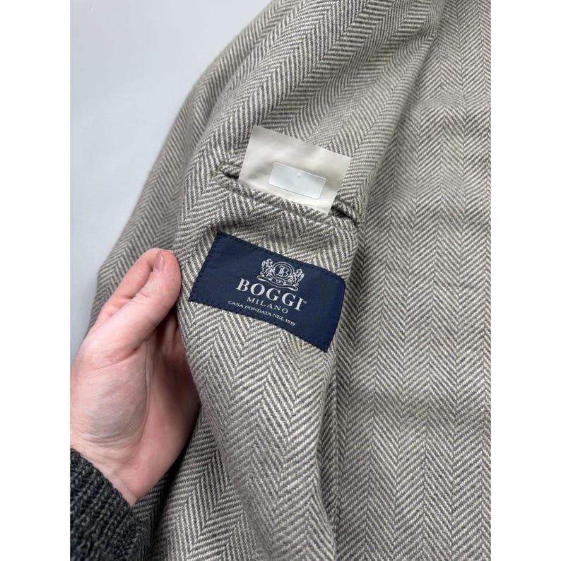 May include: A light beige and white herringbone patterned blazer featuring a dark blue interior label with the white text Boggi Milano Casa Fondata Nel 1939 and a small white rectangular tag positioned above the brand label on the inner lining.