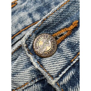 May include: Close-up of a Wrangler Authentics button on blue denim fabric. The button is a tarnished gold color with the brand name and the word "Authentics" around the edge. The denim has visible stitching.