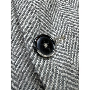 May include: A close-up view of a single dark brown, circular button with a glossy finish, centered on a grey and white fabric featuring a prominent herringbone weave pattern. The button is secured with thread and sits against the textured textile.