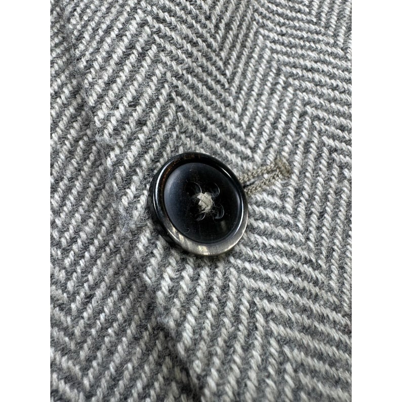 May include: A close-up view of a single dark brown, circular button with a glossy finish, centered on a grey and white fabric featuring a prominent herringbone weave pattern. The button is secured with thread and sits against the textured textile.