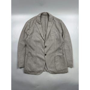 May include: A light beige blazer with a subtle micro-patterned texture, featuring a notched lapel, two dark buttons for closure, a single chest pocket, and two large patch pockets on the front. The garment has long sleeves and a structured fit.