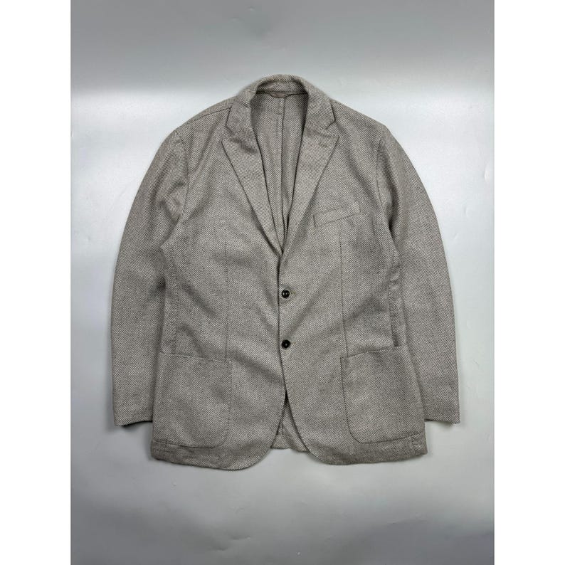 May include: A light beige blazer with a subtle micro-patterned texture, featuring a notched lapel, two dark buttons for closure, a single chest pocket, and two large patch pockets on the front. The garment has long sleeves and a structured fit.