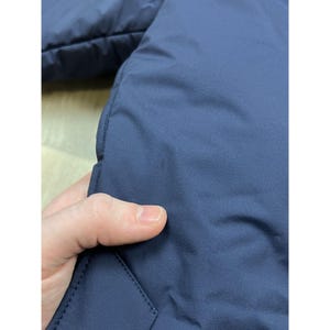 May include: Close-up of a navy blue jacket. The fabric appears smooth and slightly shiny. The image shows a portion of the jacket's exterior, with a visible seam and a hand touching the material.