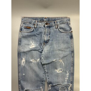 May include: Light blue denim jeans with distressed detailing, featuring multiple rips and tears across the front. The jeans have a classic five-pocket design, a button and zipper closure, and a brown leather patch on the back pocket.