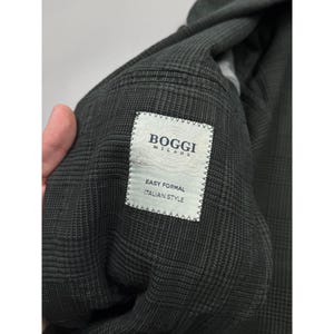 May include: A dark gray blazer with a subtle plaid pattern. A white rectangular fabric label is stitched to the interior, featuring the text BOGGI MILANO, EASY FORMAL, and ITALIAN STYLE in dark blue lettering with decorative flourishes.